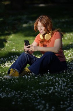 Boynunda kulaklıklarla papatya tarlasında cep telefonu kullanan gülümseyen kızıl saçlı kadın.