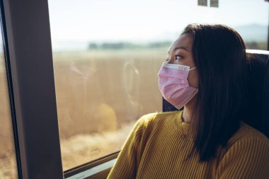 Minibüste pembe maskeli, pencereden dışarı bakan Asyalı güzel bir genç kadın var.