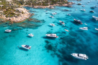 Lüks yatların ve teknelerin, seyrek bitki örtüsü olan kayalık bir sahil şeridi yakınındaki Akdeniz 'in berrak turkuaz sularında gezindiği hava manzarası. Üst dron görüntüsü. İtalya, Sardunya 'da yaz tatili