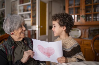 Torunu gülümseyen büyükannesine sevgi ve aile bağlarını sıcak bir ev ortamında kutlayan bir kalp çizimi gösteriyor.
