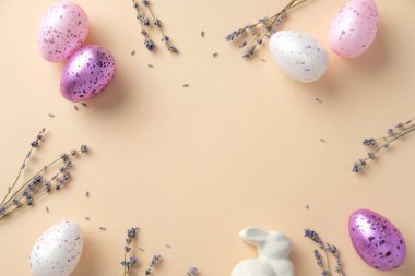 Estetik Paskalya dairesi pastel renkli benekli yumurtalar, lavanta filizi ve bej arka planda beyaz seramik bir tavşanla yatıyordu..