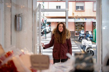 Kadın müşteri kasap dükkanına girmek için kapıyı açıyor. Elinde bir çanta dolusu et ve salam var.