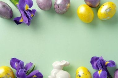 Renkli Paskalya yumurtaları ve renkli yeşil arka planda beyaz seramik tavşanı olan canlı mor irisler. Düz konum, üst görünüm, kopyalama alanı