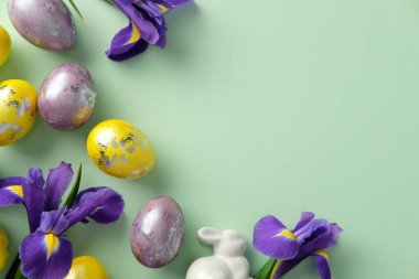Süslü yumurtalar, beyaz seramik bir tavşan ve yeşil arka planda mor süsen çiçekleri ile Pastel Paskalya düzenleme.