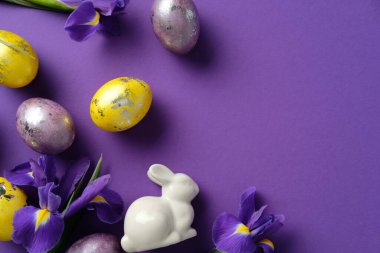 Paskalya düz kompozisyonu boyalı yumurtalar, beyaz seramik bir tavşan ve mor bir arka planda mor irisler.