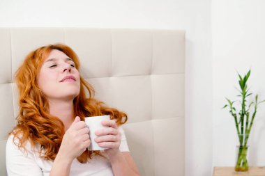 Kızıl saçlı kadın sıcak bir kahvenin tadını çıkarırken yatakta dinleniyor, huzurlu bir sabahın tadını çıkarıyor.