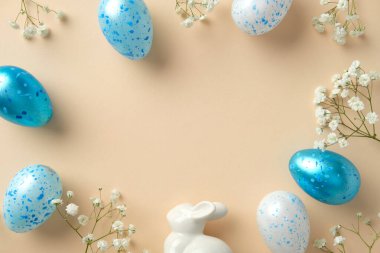 Paskalya düzlüğü mavi benekli yumurtalar, beyaz çiçekler ve bej arka planda seramik bir tavşanla yatar.