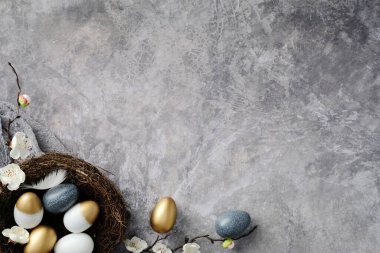 Yuvadaki zarif paskalya yumurtaları, dokulu gri arka planda beyaz çiçeklerle çevrili. Mutlu Paskalya tebrik kartı tasarımı.