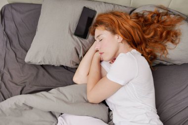 Cep telefonunun yanında uyuyan kızıl saçlı kadının üst görüntüsü.