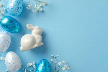 Beyaz seramik tavşan mavi ve beyaz benekli Paskalya yumurtaları ve açık mavi arka planda beyaz çiçekler arasında