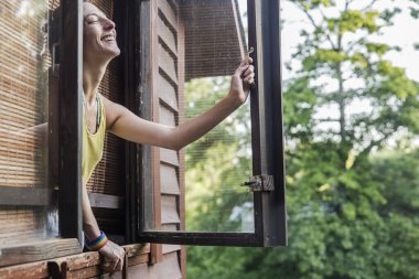 Bir kadın pencerenin önünde duruyor, gülümsüyor ve sabahın köründe temiz havayı ve yemyeşil ağaçları kucaklıyor..