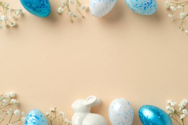 Zarif Paskalya düzlüklerinde mavi benekli yumurtalar, seramik bir tavşan ve bej arkaplanda beyaz çiçekler bulunur. Mutlu Paskalyalar.