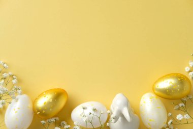 Havalı Paskalya yatağı. Beyaz ve altın benekli yumurtalar, seramik bir tavşan heykeli, ve parlak sarı bir fon üzerinde narin beyaz çiçekler.