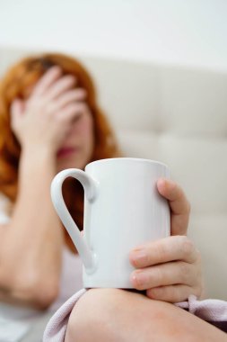 Kızıl saçlı bir kadın elinde fincanla kanepede dinlenirken şiddetli baş ağrısı çekiyor.