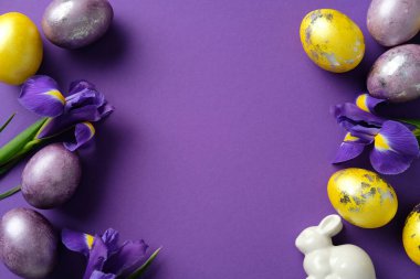 Renkli yumurtalar, canlı mor irisler ve mor arka planda beyaz seramik bir tavşan içeren Paskalya düz kompozisyonu.