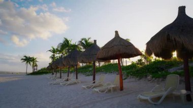 Güneşin doğuşuyla birlikte Meksika 'daki sakin sahil sahnesinde beyaz kumsalda bir sıra saman şemsiyesi ve şezlonglar, parçalı bulutlu bir gökyüzünün altında, yemyeşil palmiye ağaçlarıyla çevrili.