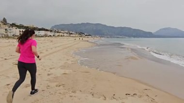 Pembe gömlekli ve siyah taytlı bir kadın bulutlu bir günde sakin deniz kenarında kumlu bir sahil boyunca koşuyor. Arkasında dağlar ve binalar var.