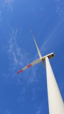 Yükselen beyaz bir rüzgar türbini açık mavi gökyüzüne karşı duruyor, temiz enerjiyi ve sürdürülebilir güç kaynaklarını simgeliyor. Uzun, parlak bıçakları rüzgarı etkili bir şekilde yakalamak için hazırdır.