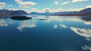 Yeni Zelanda 'nın Canterbury bölgesindeki Tekapo Gölü manzarası, berrak mavi sularını, küçük bir adayı ve bulutlarla dolu bir gökyüzünün altındaki dağlık zeminini sergiliyor..