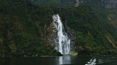 Görkemli şelale, Milford Sound, Yeni Zelanda 'daki gür, orman kayalıklarından aşağı, önplanda sakin, yansıtıcı fiyort ile doğal güzelliği ve sükuneti yakalar.
