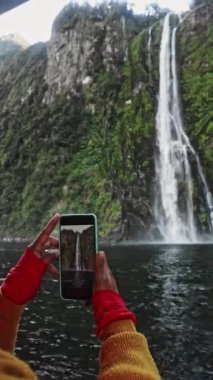 Bir kadın, Milford Sound, Yeni Zelanda 'daki yemyeşil kayalıklardan dökülen çarpıcı bir şelalenin fotoğraflarını çekiyor. Sahne, doğal güzellik ve sükunetin özünü yakalar.
