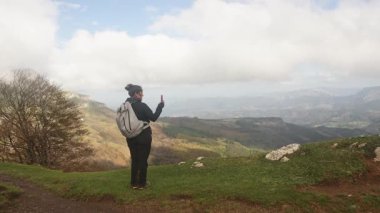 Bir kadın yürüyüşçü Gorbea Doğal Parkı 'ndaki çimenli bir dağ yamacında duruyor ve akıllı telefonuyla video çekiyor. Vadinin görkemli manzarası uzaklarda ortaya çıkıyor.