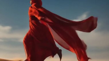 Akan kırmızı kumaşa sarılmış zarif dişi, sakin bir çöl manzarasında gösteri yapıyor. Güneş ışığı kumda dinamik gölgeler oluşturur ve hareketin akıcılığını arttırır..