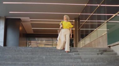 Parlak sarı bluzlu olgun bir iş kadını şehir merdivenlerinden iniyor, kendine güven ve tarz yayıyor. Bir tablet taşıyor, profesyonelliği ve amacı olduğunu gösteriyor..