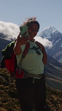 Gülümseyen bir bayan yürüyüşçü akıllı telefonuyla muhteşem arka planda selfie çeker. Yeni Zelanda 'da yemek pişirmek, açık bir günde Güney Alplerinde maceranın neşesini yakalamak.