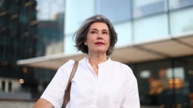 Şık beyaz kıyafetli, kendine güvenen olgun bir iş kadını, şehir ortamında profesyonelliği ve başarıyı somutlaştıran modern bir şehir zeminine karşı duruyor..