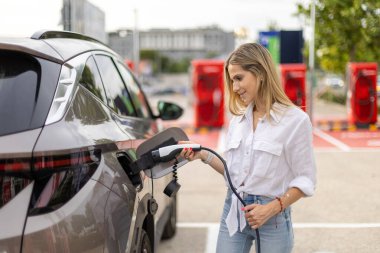 Elektrikli bir arabayı şarj ederken elinde güç kaynağı kablosu tutan iş kadını, sürdürülebilir enerji ve çevre dostu taşımanın önemini vurguluyor