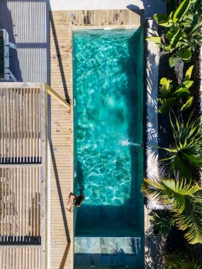 Güzel bir turkuaz havuza girmek üzere olan bir kadının hava manzarası yemyeşil bir tropikal bahçede ahşap bir güvertenin yanındaki havuza dalış havuzu.