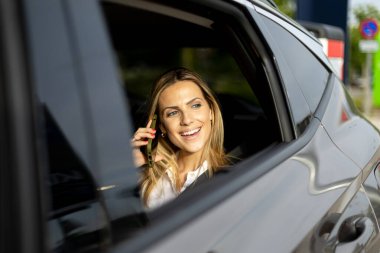 Bir şarj istasyonunda elektrikli arabasında otururken gülümseyen iş kadını telefonla konuşuyor.