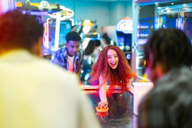 Neşeli genç bir kadın arkadaşlarıyla hava hokeyi oynuyor, oyun salonunda eğleniyorlar.