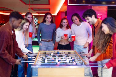 Bir grup arkadaş renkli bir barda masa futbolu oynayıp içki içerek eğleniyorlar.