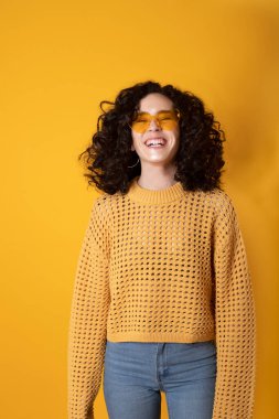 Kıvırcık siyah saçlı ve sarı arka planda moda gözlüklü gülen kız.