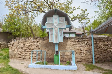 Geleneksel kırsal yol kenarı haçı, mavi boyanmış ahşap haç ve metal tepe örtüsü, bir köyde taş bir duvara dayanıyor.