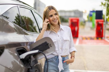 Gülümseyen iş kadını, şarj istasyonunda telefonla konuşurken elektrikli arabasını şarj ediyor.
