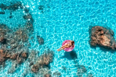 Yazın günbatımında mavi denizde pembe yüzme yüzüğüyle yüzen genç bir kadının hava manzarası. Kızlı tropik manzara, temiz su, taşlar, kayalar, kumlu plaj. Üst Manzara. Lefkada Adası, Yunanistan