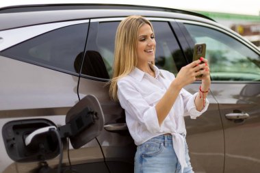 Elektrikli arabası istasyonda şarj olurken akıllı telefon kullanan gülümseyen iş kadını sürdürülebilir bir yaşam tarzının keyfini çıkarıyor.