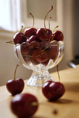 Taze toplanmış kirazlar cam bir kaseden ahşap bir masaya dökülüyor, canlı ve iştah açıcı bir görüntü yaratıyor.