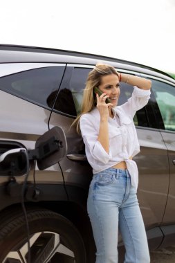 Modern bir şarj istasyonunda elektrikli arabasını şarj ederken akıllı telefonuyla konuşan gülümseyen bir iş kadını.