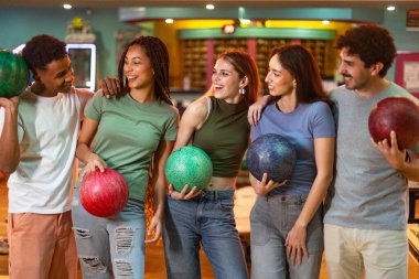 Beş genç arkadaş bovling toplarını tutarak ve gülümseyerek birlikte eğleniyorlar.