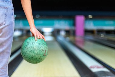 Yeşil bir bowling topu tutan kadın, bovling oynamaya hazırlanıyor, arka planda bulanık bowling şeritleri var.