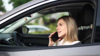 Sarışın profesyonel iş kadını, gündüz vakti şehir içi banliyö yolunda güvenli bir şekilde modern aracı kullanırken cep telefonuyla neşeli bir şekilde konuşuyor.
