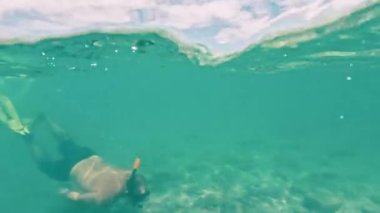 Ekranda, küçük bir volkanik adanın yakınında turkuaz sığ suda şnorkelle yüzen bir adam görülüyor.