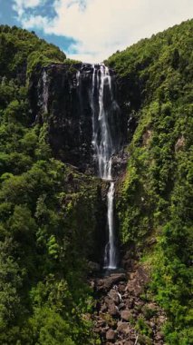 Yeni Zelanda 'nın el değmemiş doğal ortamında berrak mavi bir gökyüzünün altında canlı yeşil yapraklarla çevrili engebeli uçurumlardan nefes kesici bir şelale düşüyor.