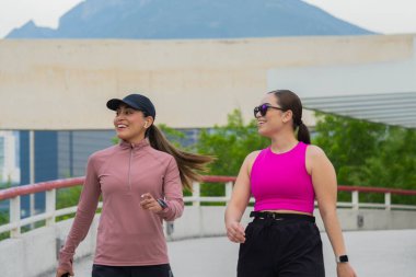 Spor kıyafetleri içinde iki Latin kadın. Gülümsüyor ve açık bir yolda birlikte yürüyorlar. Keyifli bir aktivitenin tadını çıkarıyorlar.
