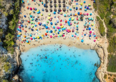 Kalabalık kumlu sahillerde renkli şemsiyeler, mavi denizde yüzücüler, güneşli yaz günlerinde yeşil ağaçlar. Mallorca, Balear Adaları, İspanya. Tropik insansız hava aracı görüntüsü. Turkuaz su. Seyahat