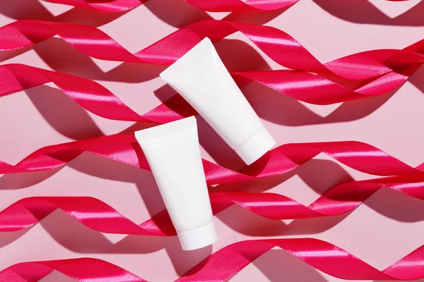 Blank white plastic cosmetic tubes with pink ribbons on pink background. Cosmetic beauty product branding mockup. Copy space